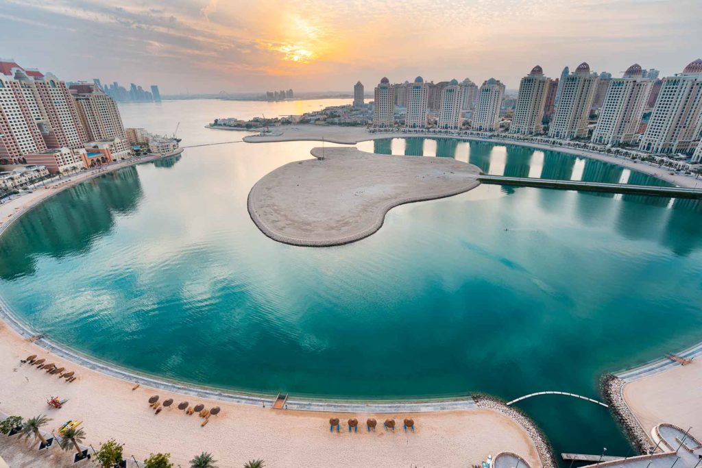Vista panorâmica do the pearl em Doha com prédios altos rodeando uma lagoa com uma ilha no meio e o sol ao fundo