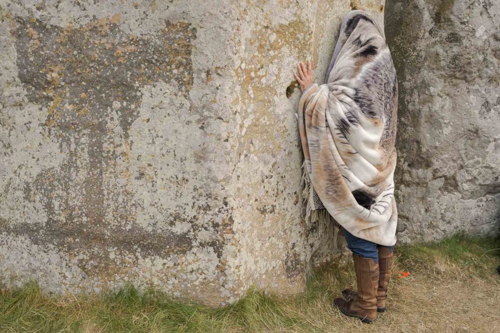 Mulher coberta por um pano encostada na pedra de Stonehenge da cabeça aos pés