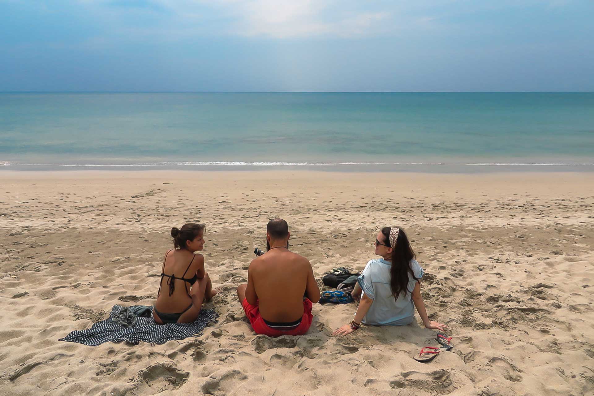 Tiago playing ukulele in the middle of Fernanda and a friend alone in the beach with a blue sky when we were backpacking in Thailand