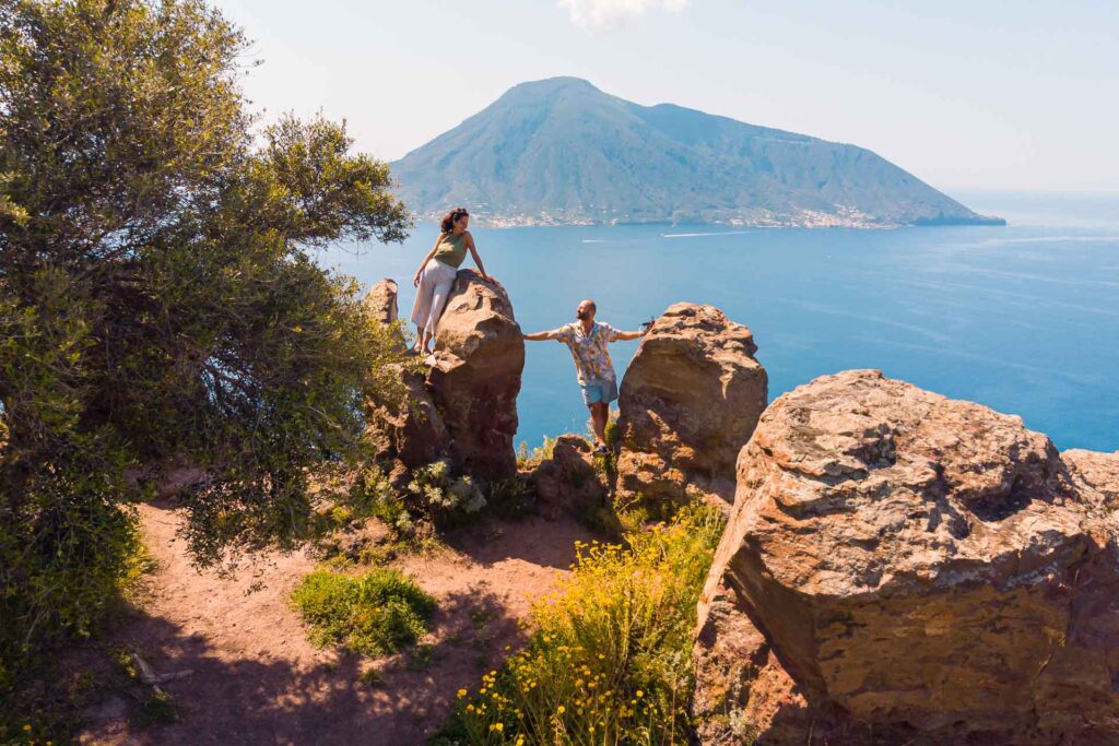 Tiago and Fernanda on a cliff in Lipari Islands with the view of an island in the background