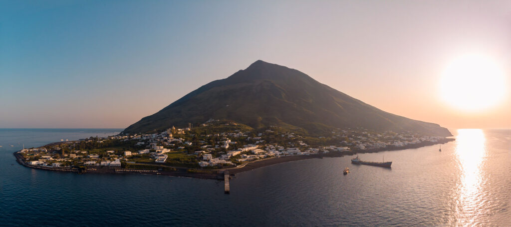 Vista da ilha de Stromboli com um vulcão no meio da ilha, o sol se pondo no lado direito e a cidade no lado esquerdo da ilha