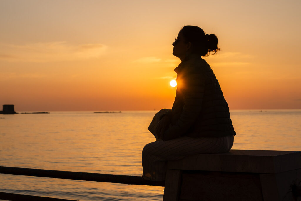 A silhueta da Fernanda sentada na beira do mar vendo o por do sol