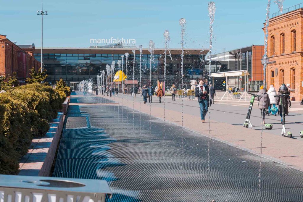 Uma grande praça no centro de Lodz na Polônia with uma fonte dançante no caminho e o Manufaktura ao fundo