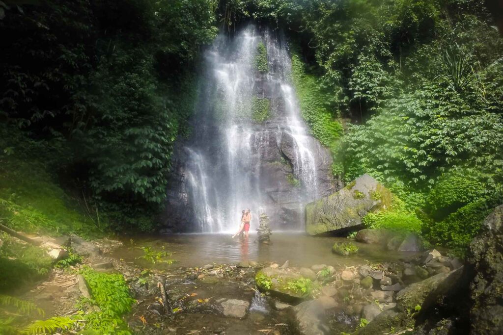 A cachoeira Munduk caindo em um lago bem raso com a Fernanda e Tiago embaixo da cachoeira