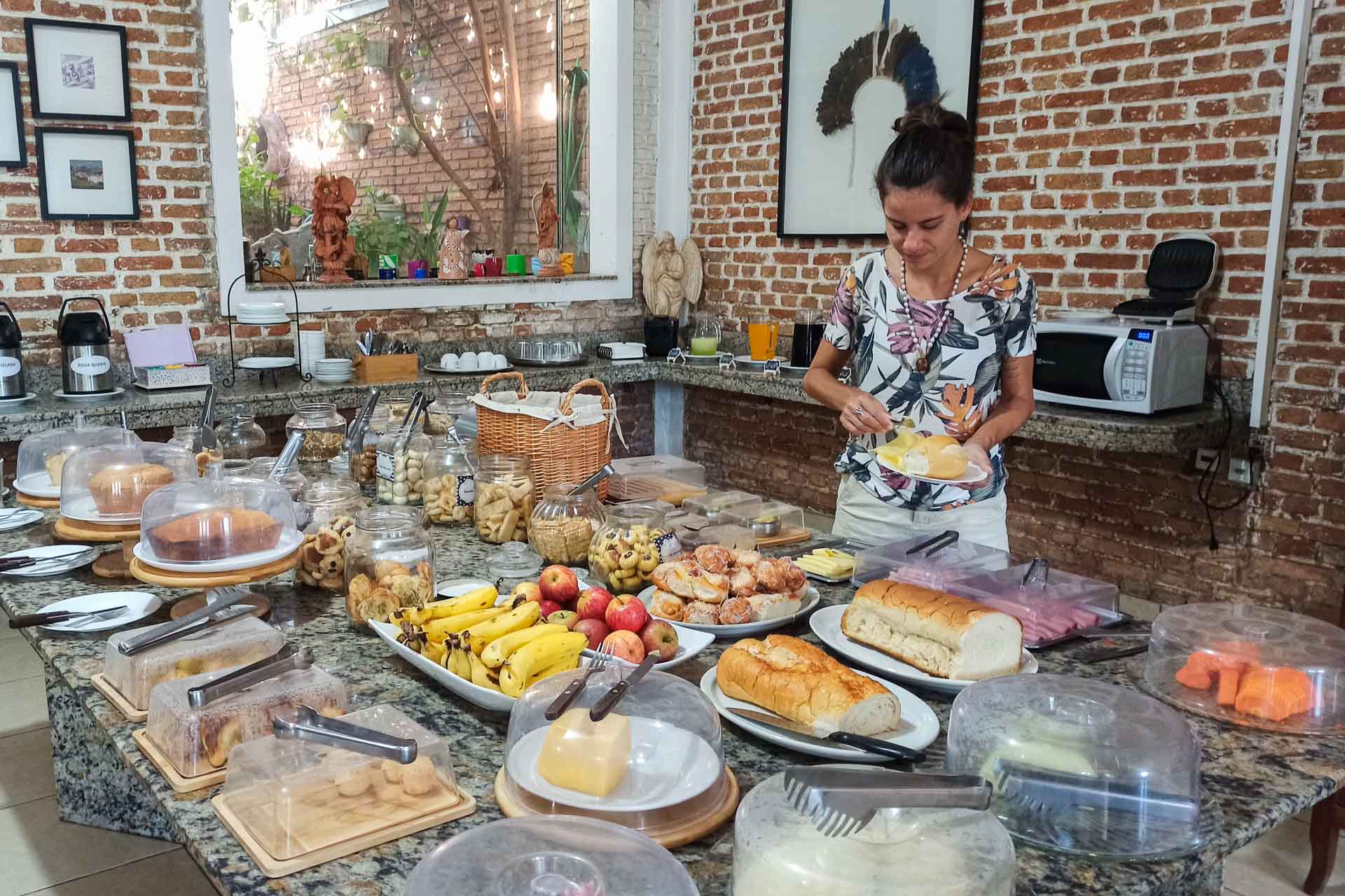 Fernanda in front of a large table of breakfast full of food, fruit and juices, in a room with brick walls at the Hotel Virginia in Corumba, Brazil